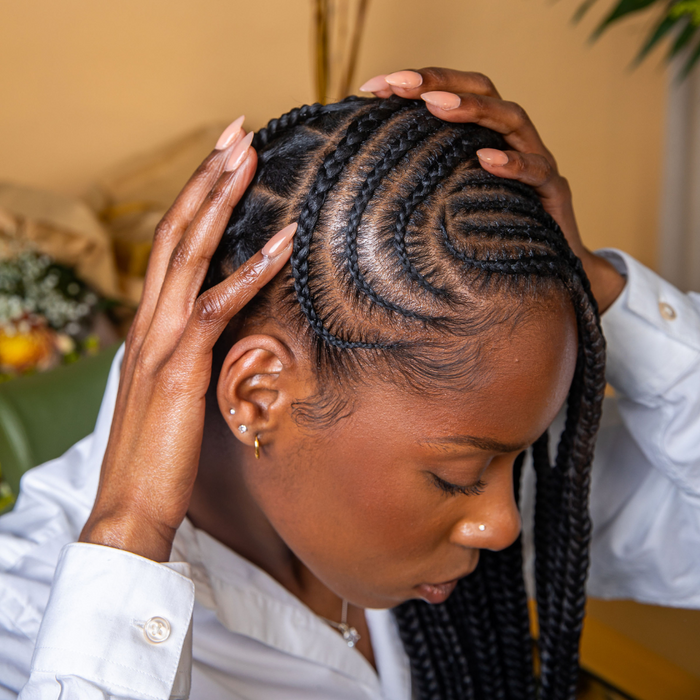 Person with braided hair feeling her braided hair while wearing a white shirt against a beige background