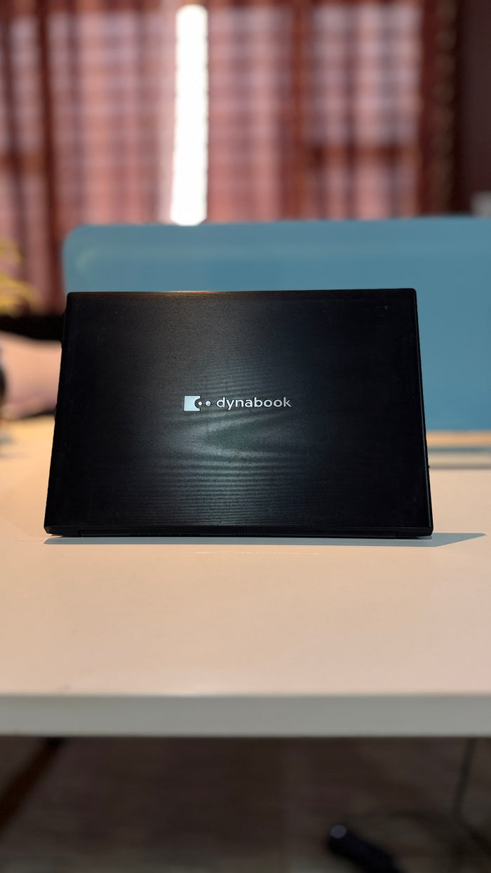 A black Dynabook laptop closed and standing upright on a white table, with a blurred indoor background featuring a blue chair and warm-toned curtains.