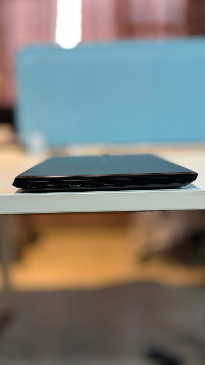 A black slim laptop shown from the side, resting on a white table with a blurred indoor background.